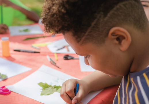Child drawing leaves at outdoor art table.